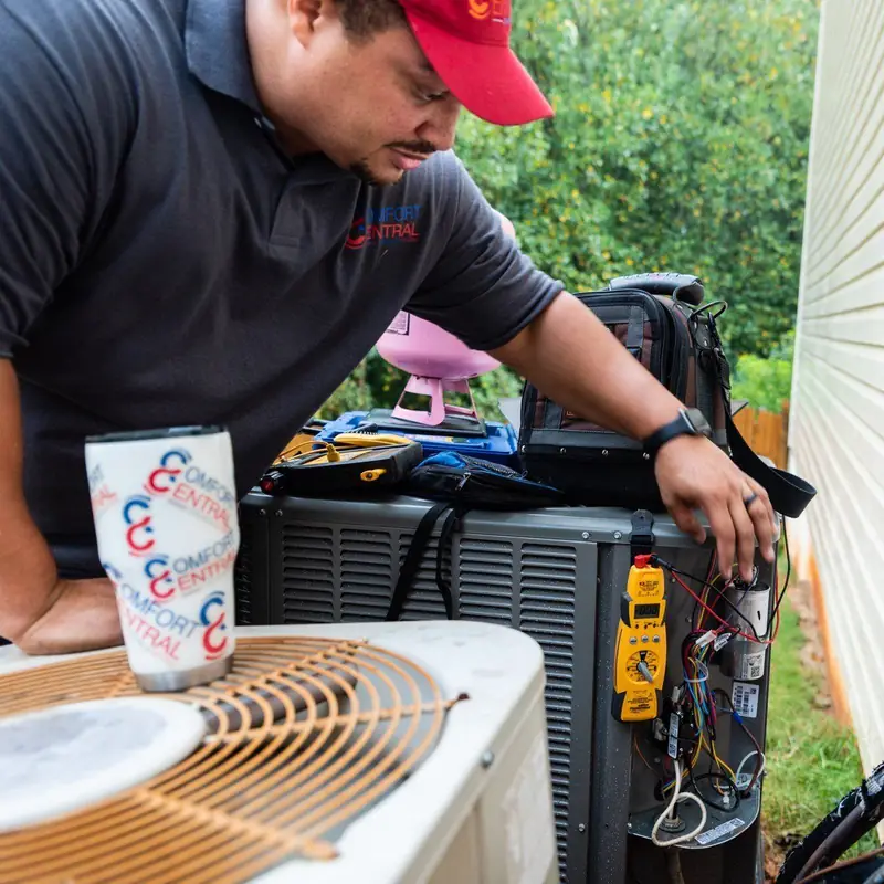 A Comfort Central technician in a branded polo leaning into an open condenser electrical panel with a meter clipped to the capacitor wiring
